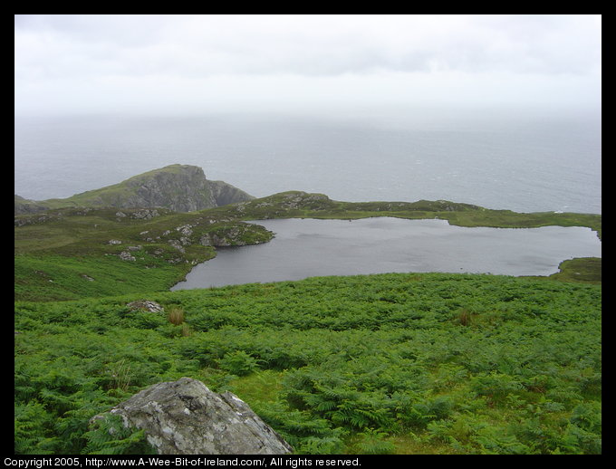 Loch O'Mulligan Slieve League. Far below there is a lake reflecting the sky and beyond the lake are a rocky hill and the ocean.