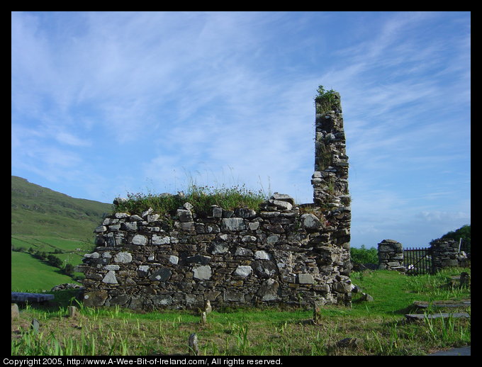 This a view of the side of the ruin Saint Cartha's Church near Kilcar, Donegal. The gate to the churchyard and stone wall are in the background. In the foreground are very weathered stone grave markers. The older markers are standing while the newer ones lie flat.