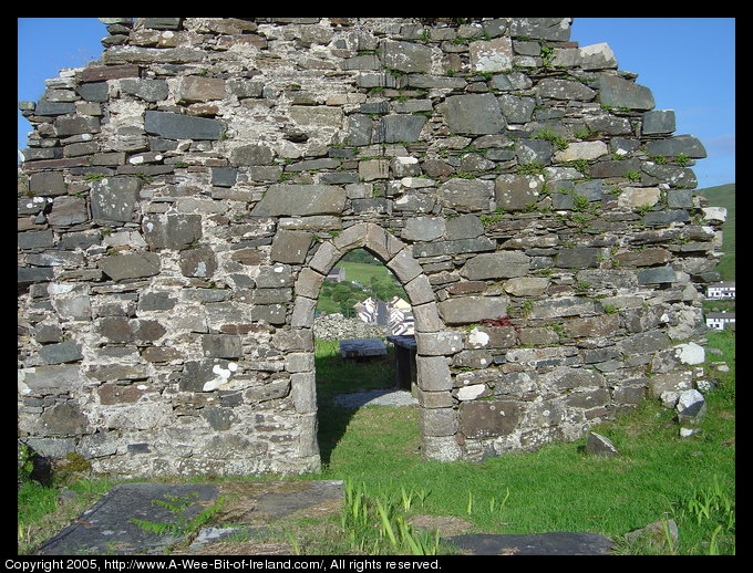Saint Cartha's Church near Kilcar, Donegal with Kilcar Village visible through the arch. In the foreground are some flat slab grave markers and iris plants that are not blooming.